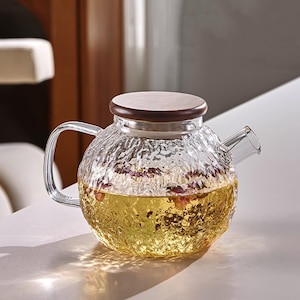 May include: A clear glass teapot with a textured surface, filled with golden liquid and rose petals. It has a wooden lid and a curved glass handle. The teapot is designed for brewing tea.