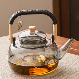 Puede incluir: Una tetera de vidrio transparente con un asa negra y detalles de madera. La tetera tiene una tapa de vidrio y un pico. El interior está lleno de té. La tetera está decorada con diseños de pan de oro y pequeños amuletos colgantes.