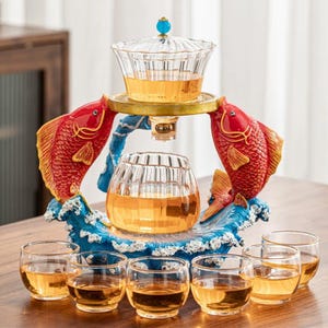 May include: A decorative tea set featuring a glass teapot and six small glasses. The set is supported by a blue and gold base with two red fish sculptures. The teapot is filled with tea, and the glasses are partially filled.