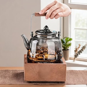 Könnte beinhalten: Eine Teekanne aus klarem Glas mit dunkelgrauer Tönung, ausgestattet mit einem schwarzen Deckel mit spitzem Knauf, einem geschwungenen Ausguss und einem Metallgriff mit Holzeinsatz. Die Teekanne steht auf einem quadratischen Holzsockel.