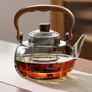 Puede incluir: Una tetera de vidrio transparente con asa y tapa de madera. La tetera tiene un infusor incorporado y está llena de té. El asa es curvada y de madera de color marrón claro.