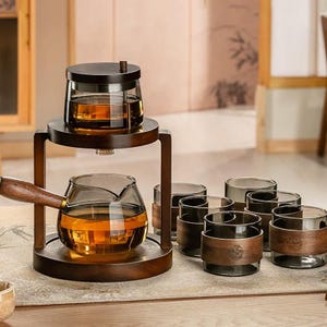 Puede incluir: Un juego de té de madera con una tetera de vidrio, un infusor de vidrio y seis vasos a juego. La tetera tiene un asa de madera. El juego está lleno de té de color ámbar. Los vasos tienen bandas de madera alrededor. El juego está sobre un soporte de madera.