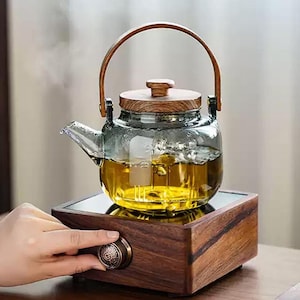 Peut inclure: Une théière en verre avec une poignée et un couvercle en bois, remplie de thé, est posée sur une base en bois. La théière est transparente, permettant de voir la couleur du thé. Une main tourne un bouton sur la base en bois. La théière mesure environ 15 cm de haut.