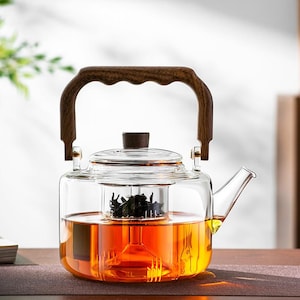 Può includere: Una teiera in vetro trasparente con manico e coperchio in legno. La teiera è riempita di tè color ambra e ha un infusore incorporato contenente foglie di tè sfuse. Il manico è curvo e in legno scuro.