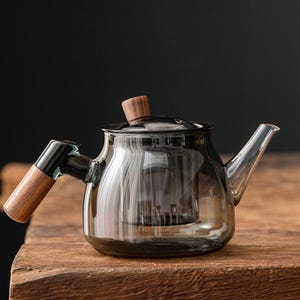 Può includere: Una teiera in vetro trasparente con una tinta scura, con manico e coperchio in legno. La teiera ha un beccuccio e un infusore integrato. Il design è moderno e minimalista, adatto per preparare il tè.