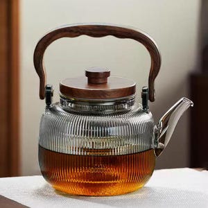 May include: A clear glass teapot with a wooden handle and lid. The teapot has vertical ridges and is filled with a golden-brown liquid. The handle and lid are made of dark wood. The spout is made of shiny metal.