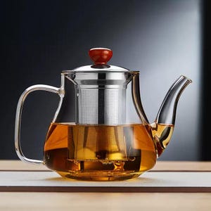 May include: A clear glass teapot with a stainless steel infuser and a red wooden knob on the lid. The teapot is filled with amber-colored tea. The handle and spout are curved. The teapot is sitting on a light-colored surface.