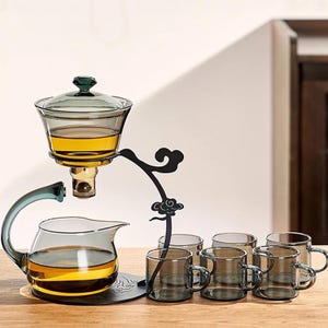 Könnte beinhalten: Ein Teegeschirr aus Glas mit einem schwarzen Metallständer. Das Set enthält eine Teekanne, einen Filter und sechs kleine Teetassen. Die Teekanne und der Filter sind mit Tee gefüllt. Das Set ist zum Brühen und Servieren von Tee konzipiert.