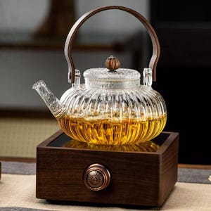 Puede incluir: Una tetera de vidrio transparente con un diseño acanalado, llena de líquido ámbar, se encuentra sobre una base de madera. La tetera tiene un asa de madera y una tapa decorativa. La base tiene un pomo color cobre. Un elegante juego de té.