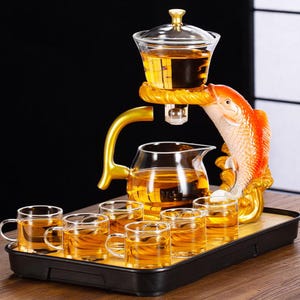 Può includere: Servizio da tè decorativo con una teiera in vetro, sei tazzine e un supporto a forma di pesce. Il supporto è dorato e arancione, con un infusore per tè in vetro. Il tè è color ambra e il set è su un vassoio nero.