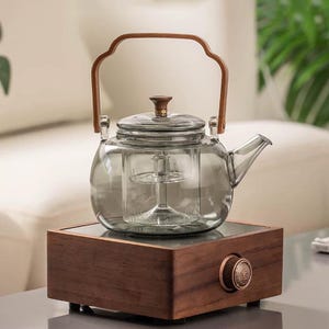 May include: A clear glass teapot with a wooden handle and lid sits atop a dark wooden base. The teapot has a built-in infuser and a spout for pouring. The base has a decorative knob. The teapot is designed for brewing tea.