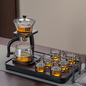 Puede incluir: Un juego de té de vidrio con base y soporte de madera. El juego incluye una tetera, un recipiente para preparar y seis tazas pequeñas, todas llenas de té color ámbar. Los elementos de madera son de color marrón oscuro.