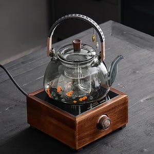 May include: A clear glass teapot with a black handle and wooden accents sits on a wooden base. The teapot has a floral design and a spout. The base has a control knob and a black surface.