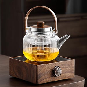 Puede incluir: Una tetera de vidrio transparente con asa y tapa de madera, llena de líquido amarillo, se encuentra sobre una base de madera con un mando de control. La tetera tiene un diseño acanalado y un borde de acero inoxidable.