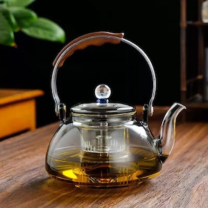 Op de afbeelding: Een transparante glazen theepot met een houten handvat en een roestvrijstalen infuser. De theepot is gevuld met een goudkleurige vloeistof, waarschijnlijk thee, en staat op een houten oppervlak. Het deksel heeft een kleine, transparante knop met een blauwe basis.