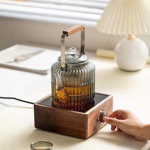 Może przedstawiać: Szklany czajnik z drewnianym uchwytem i metalową ramą, wypełniony herbatą. Czajnik stoi na drewnianej podstawie z czarną powierzchnią grzewczą. Ręka obraca drewniane pokrętło na podstawie. Czajnik stoi blisko okna i lampy.