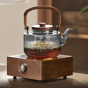 May include: A glass teapot with a wooden handle and lid sits on a wooden base with a control knob. The teapot has a ribbed design and is filled with a dark liquid, likely tea. The base appears to have a heating element.