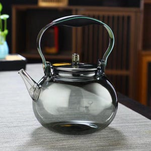 May include: A clear glass teapot with a rounded body and a curved metal handle. The spout is angled, and the lid is topped with a decorative knob. The teapot is designed for brewing tea and has a capacity of approximately 1 litre.