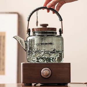 May include: A glass teapot with a wooden lid and handle, filled with water. The teapot is set on a wooden base with a copper-coloured button. The handle is wrapped in a dark material. A small decorative charm hangs from the handle.