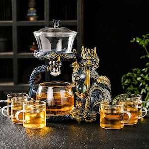 以下が含まれることがあります： ガラスのティーポットと6つのガラスのティーカップを備えた、装飾的なティーセット。 青と金のドラゴンの彫刻がセットを支えています。 お茶は琥珀色で、セットは暗い表面に配置されています。