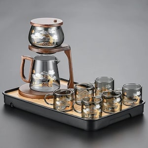 Puede incluir: Un juego de té con acabado gris oscuro, que incluye una tetera, un recipiente con tapa y seis tazas a juego. Cada pieza está decorada con un diseño de árbol dorado y plateado. El juego se presenta en una bandeja negra con una inserción de bambú.