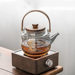 May include: A clear glass teapot with a wooden handle and lid, featuring a gold koi fish design. The teapot sits on a wooden base with a control knob. The tea inside is amber-colored.