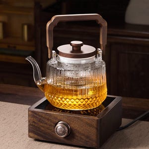 May include: A glass teapot with a wooden handle and lid, filled with amber liquid, sits on a wooden base with a control knob. The teapot has a textured, patterned design. The base has a dark brown finish.