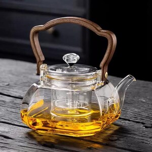 Op de afbeelding: Een heldere glazen theepot met een houten handvat en een glazen infuser. De theepot is gevuld met een goudkleurige vloeistof. De handgreep is gebogen en gemaakt van lichtbruin hout. Het deksel heeft een kristalachtige knop.