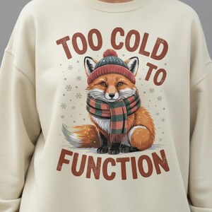 Puede incluir: Sudadera color crema con un zorro con una bufanda y gorro a cuadros rojos y verdes. El texto "TOO COLD TO FUNCTION" está impreso encima y debajo del zorro.
