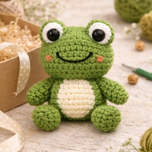 May include: A handmade green crochet frog with a cream-colored belly. The frog has large black eyes, a smiling mouth, and small pink cheeks. The frog is a soft toy, perfect for a child's room or as a decorative item.
