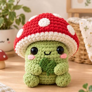 May include: A crocheted plush toy shaped like a green creature with a large red mushroom cap featuring white spots. The creature has black eyes, pink cheeks, and a small smile, and it is holding a small green four-leaf clover in its hands.