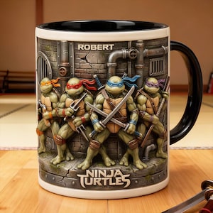 Può includere: Tazza in ceramica bianca con le Tartarughe Ninja. La tazza ha un manico e un bordo neri, con un rilievo dettagliato delle tartarughe in azione. Il nome "ROBERT" è stampato sopra le tartarughe e le parole "NINJA TURTLES" sono alla base.