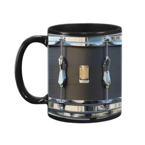 May include: Black coffee mug featuring a realistic drum design. The mug has a black handle and rim, with a detailed image of a snare drum on the front. Silver-colored hardware and a gold-colored plate are also visible.