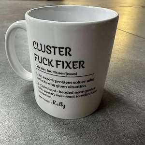 Puede incluir: Taza de cerámica blanca con el texto negro "CLUSTER FUCK FIXER". La taza incluye una definición del término, que describe a un experto en la resolución de problemas. El texto también incluye "See also: Kelly".
