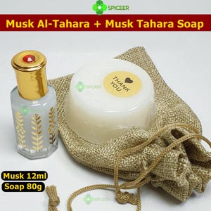 May include: A set of personal care products including a small perfume bottle, a bar of soap, and a drawstring bag. The perfume bottle is gold and clear, with a red gem on top. The soap is white with a "Thank You" sticker. The bag is beige. The text reads "Musk Al-Tahara + Musk Tahara Soap".