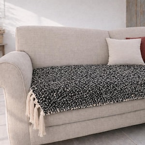 May include: A neutral-toned sofa with a black and white speckled throw and two cushions. One cushion is white, the other is a reddish-brown. The throw has a fringed edge.