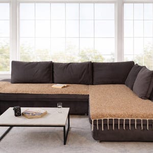 May include: A dark brown corner sofa with matching cushions and a beige throw. A white marble coffee table with a black metal frame holds a camera, a decorative box, and a glass. The sofa is in front of a large window.