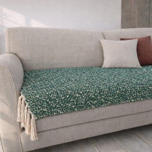 May include: A green and white textured throw blanket with tassels is draped over a light grey sofa. Two throw cushions in neutral tones are arranged on the sofa. The blanket adds colour and texture to the neutral-toned furniture.