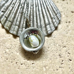 May include: A silver-toned pendant necklace featuring a small, round charm with the word "wish" and a small, white and yellow stone inside. The pendant hangs from a delicate chain, set against a seashell.