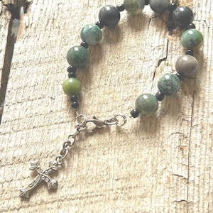 May include: A beaded bracelet featuring a mix of green, gray, and black round beads. The bracelet includes a silver-colored cross charm and a clasp closure. The beads are strung together with black beads.