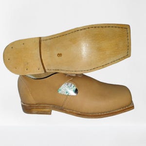 May include: A pair of tan leather shoes with a light brown sole. The sole of one shoe is visible above the other. The shoes have a classic design with a rounded toe and a low heel. The sole is marked with the number 8.