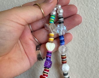 Puede incluir: Un llavero de cuentas de colores con un cierre plateado. Las cuentas incluyen perlas blancas, transparentes y de varios colores, incluyendo una rosa roja, un corazón blanco y una flor transparente. El llavero se sostiene en una mano.
