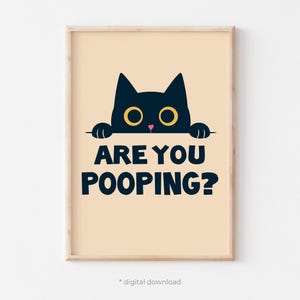 Op de afbeelding: Een ingelijste print met een zwarte kat met gele ogen en de tekst "ARE YOU POOPING?" in marineblauw. De kat kijkt over een horizontale lijn. De achtergrond is licht perzik van kleur.