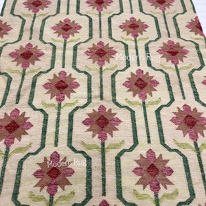 May include: A beige rug featuring a repeating floral design. The flowers are burgundy and brown, framed by a green geometric pattern. The text "Modern Rug" is displayed on the rug.