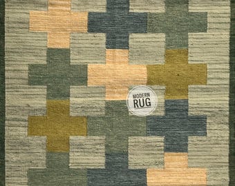 Puede incluir: Una alfombra rectangular con un patrón de cruces repetidas en tonos verdes, azules y beige. La alfombra tiene un aspecto texturizado y un borde verde oscuro. El texto "MODERN RUG" está en un círculo blanco.