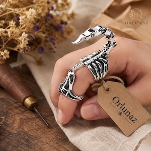 Può includere: Un anello articolato color argento a forma di scorpione, caratterizzato da una coda segmentata che si curva sopra il dito e da un corpo dettagliato. Un piccolo cartellino rettangolare in carta marrone con il testo stampato Orlunaz è attaccato all'anello.