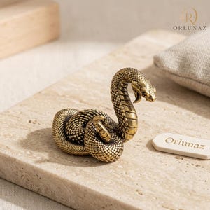 Puede incluir: Una pequeña figura de serpiente de color latón enroscada con escamas detalladas y la boca abierta. La serpiente está en una pose llamativa con la cabeza levantada. Una pequeña etiqueta rectangular blanca con el texto Orlunaz está colocada cerca.