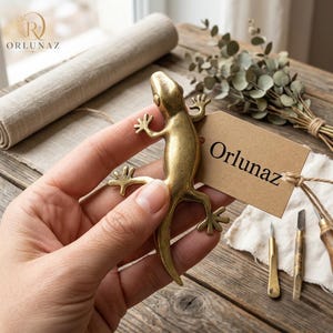 May include: A metallic gold-toned lizard figurine with a smooth, polished finish. Attached to the lizard is a rectangular brown paper tag featuring the printed text Orlunaz, secured with a thin piece of twine tied around the lizard's midsection.