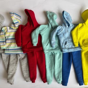 May include: Six miniature hoodie and tracksuit sets in a variety of colours. The sets include grey, red, mint green, light blue, and yellow. One set features a striped pattern. Each set has a hood and a front pocket, suitable for dolls.