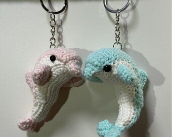 May include: Two handmade crochet dolphin keyrings. One dolphin is pink and white, the other is blue and white. Both have black eyes and silver key rings. The keyrings are hanging against a white background.